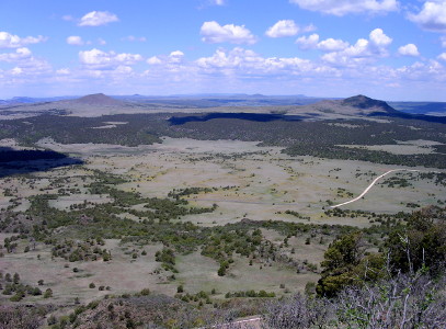 [Mountains in distance skyline with puffy white clouds above and tree dots on nearby plains along with a white ribbon that appears to be a defined road.]